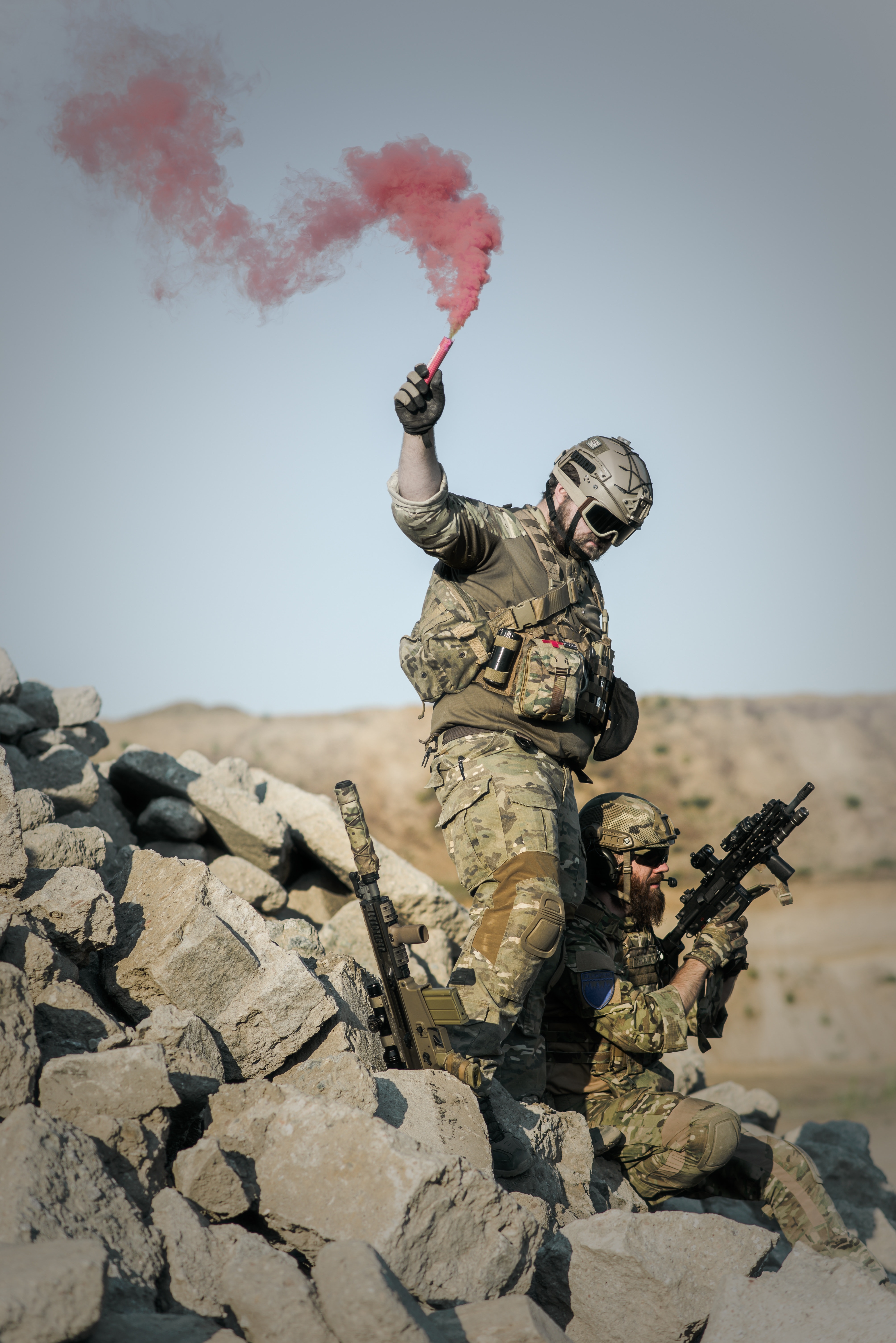 Soldier with guns on grey pile of rocks holding smoke stick during daytime