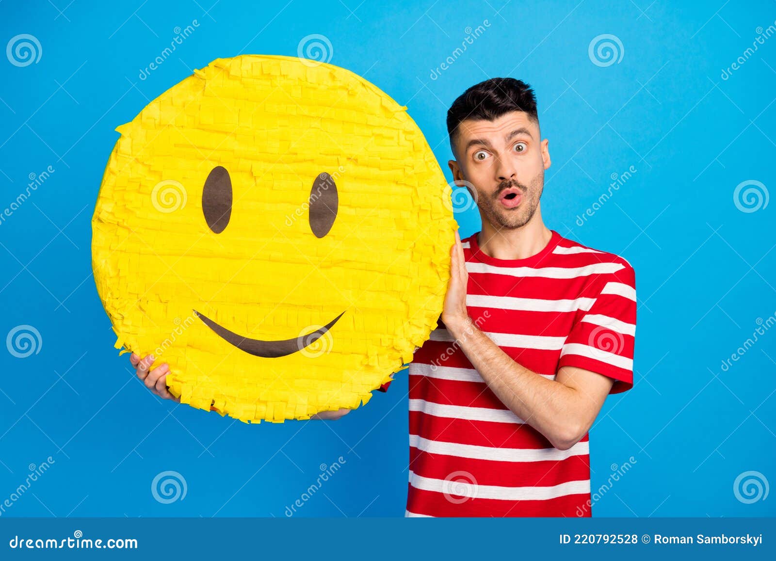 Photo of amazed shocked young man hold big paper pinata smile emoji face isolated on blue color background stock photo